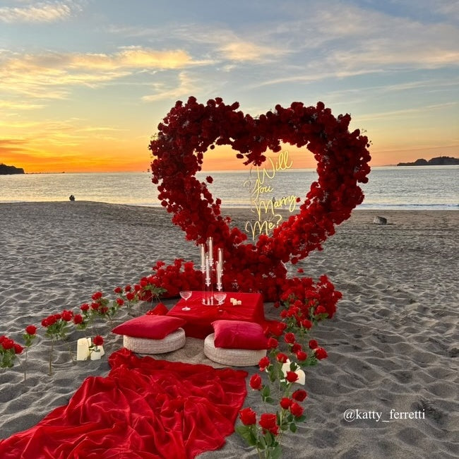 Beach proposal with red heart arch and lights saying "Will you marry me".