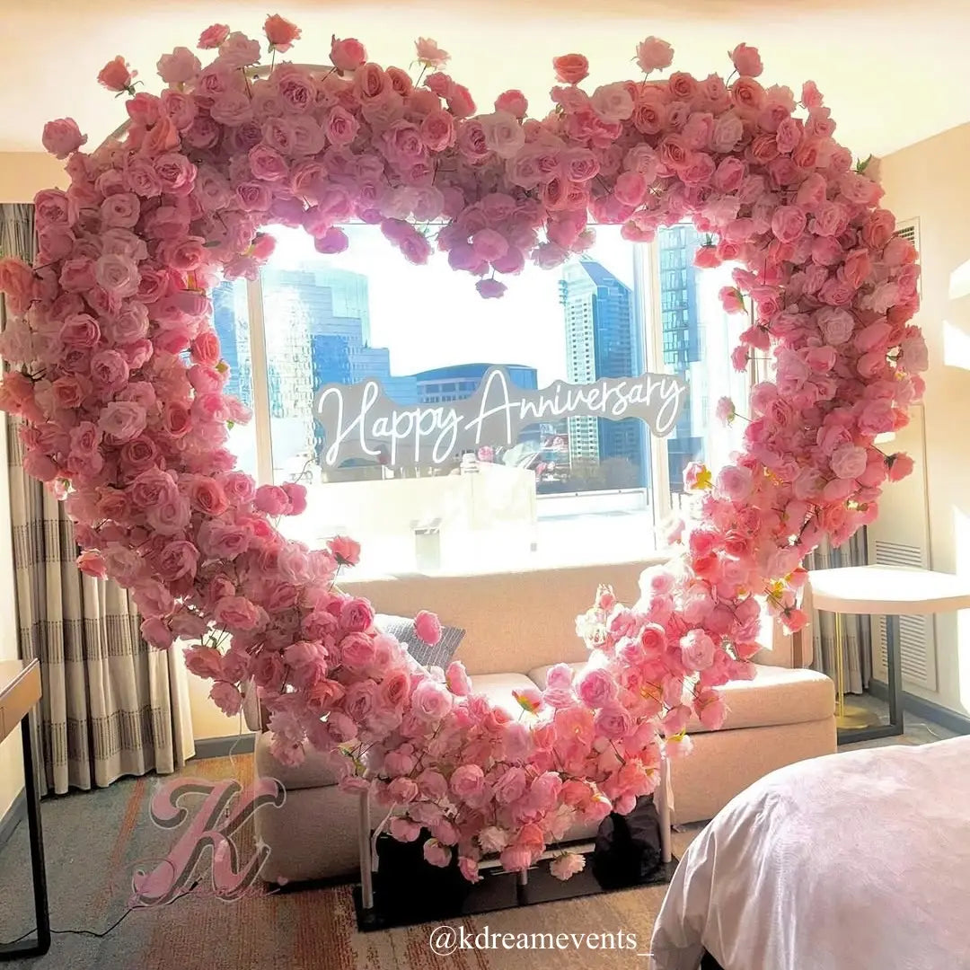 Pink heart-shaped flower arch with happy anniversary neon on bedroom.